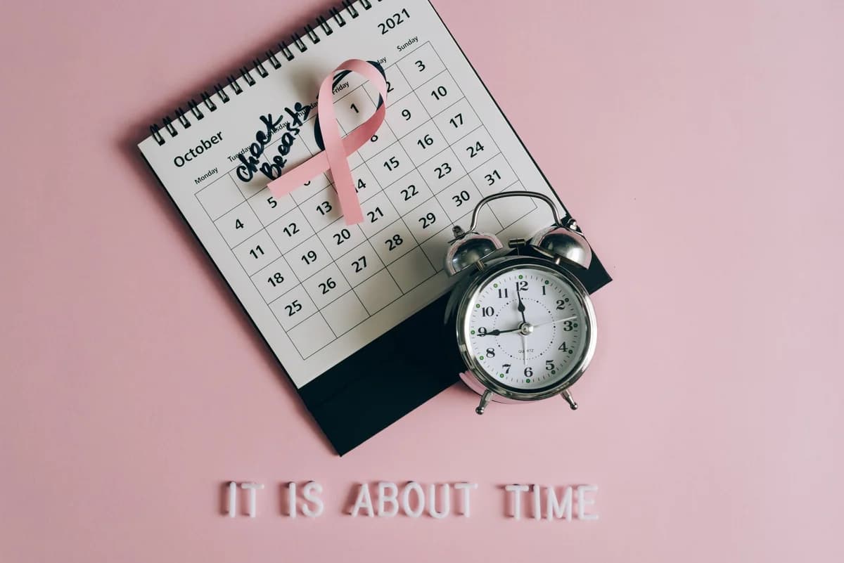 Calendar with pink ribbon, an alarm clock, and the text 'Check breasts' signifies Breast Cancer Awareness.
