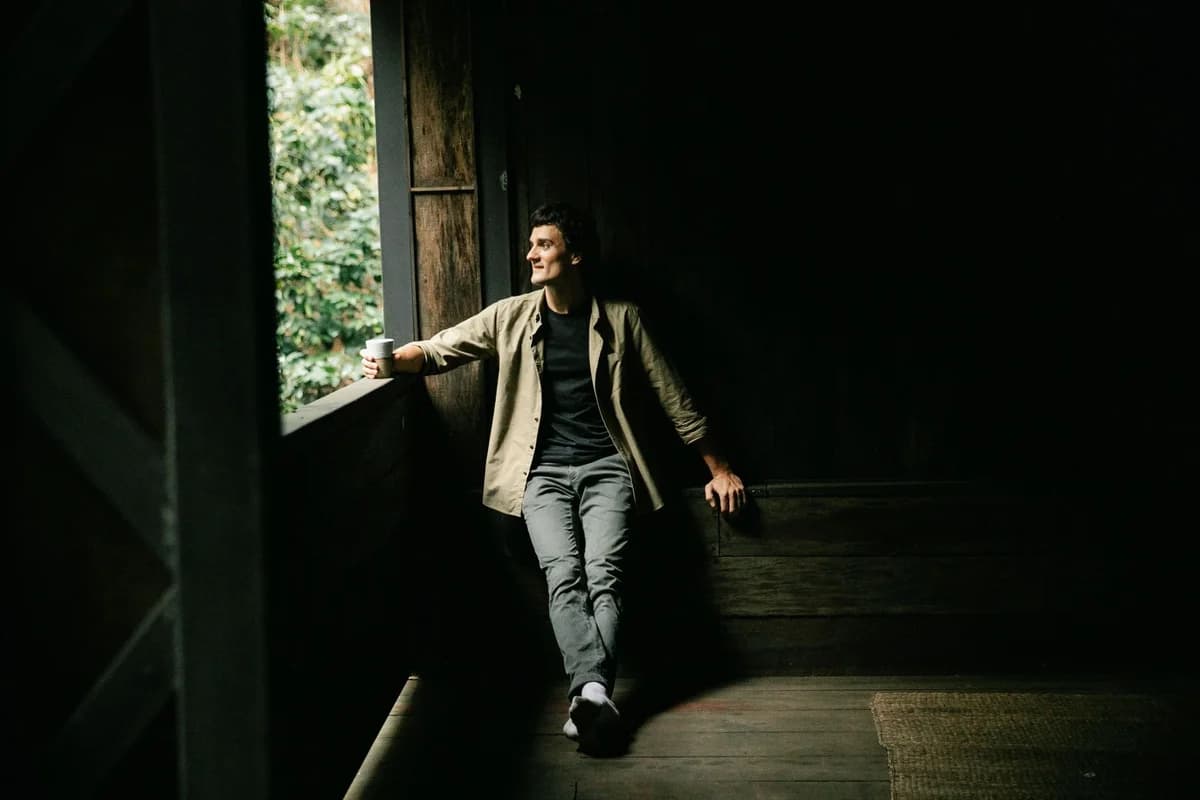 Full body of positive male looking out window while sitting in shabby wooden cabin with coffee grinder in hand in countr