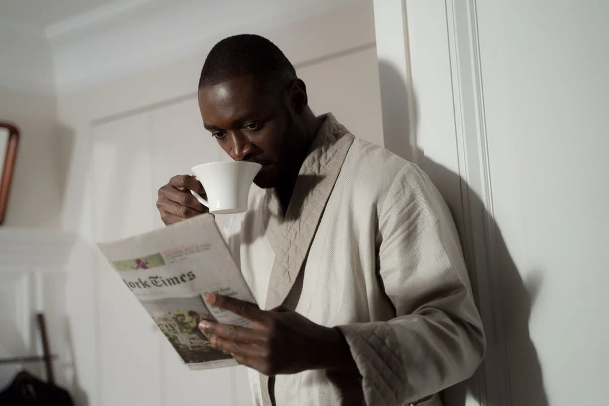 Man in a bathrobe enjoys coffee and reads a newspaper indoors.