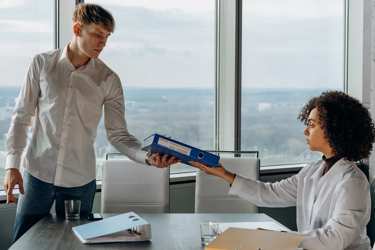 Two colleagues exchange documents in a contemporary office with a scenic city view.