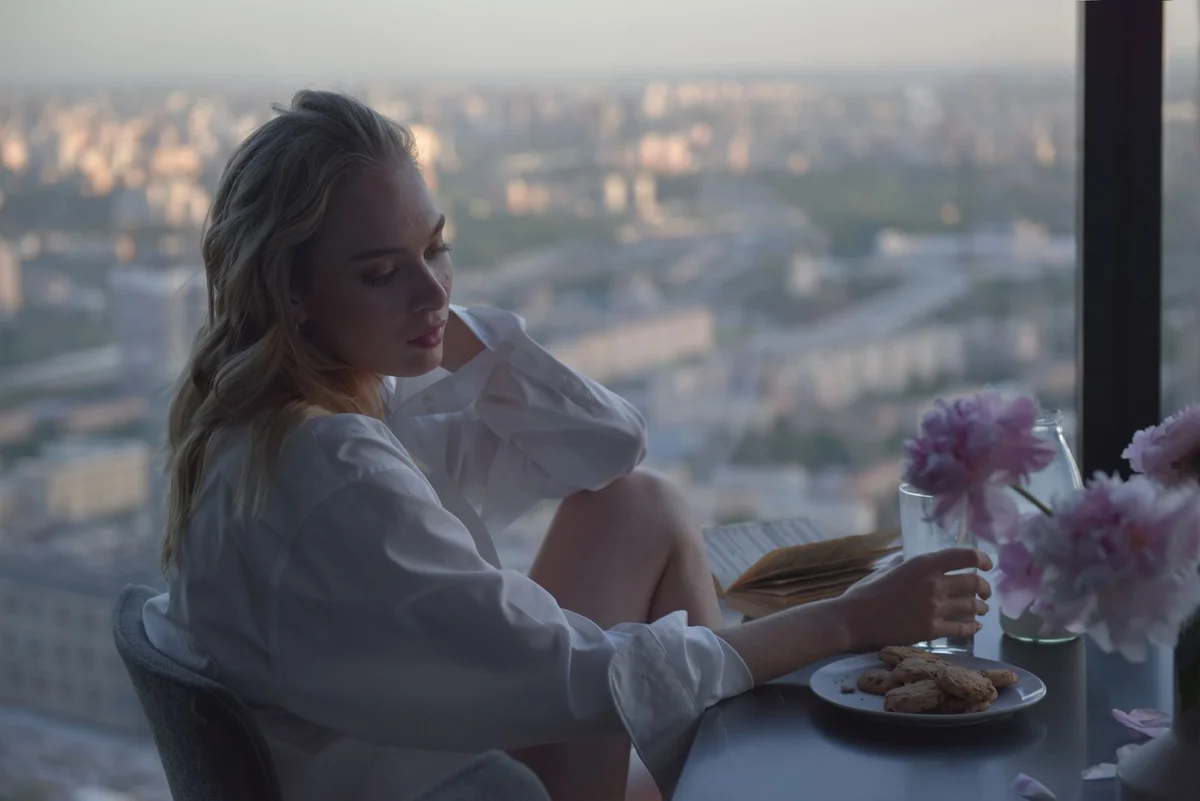 A woman in white long sleeves enjoying cookies by a city view window.