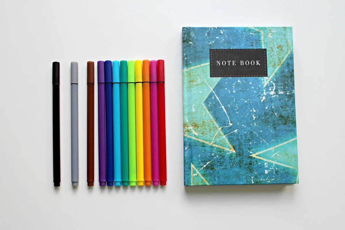 A colorful assortment of pens lined up beside a blue notebook on a white background, perfect for creative projects.