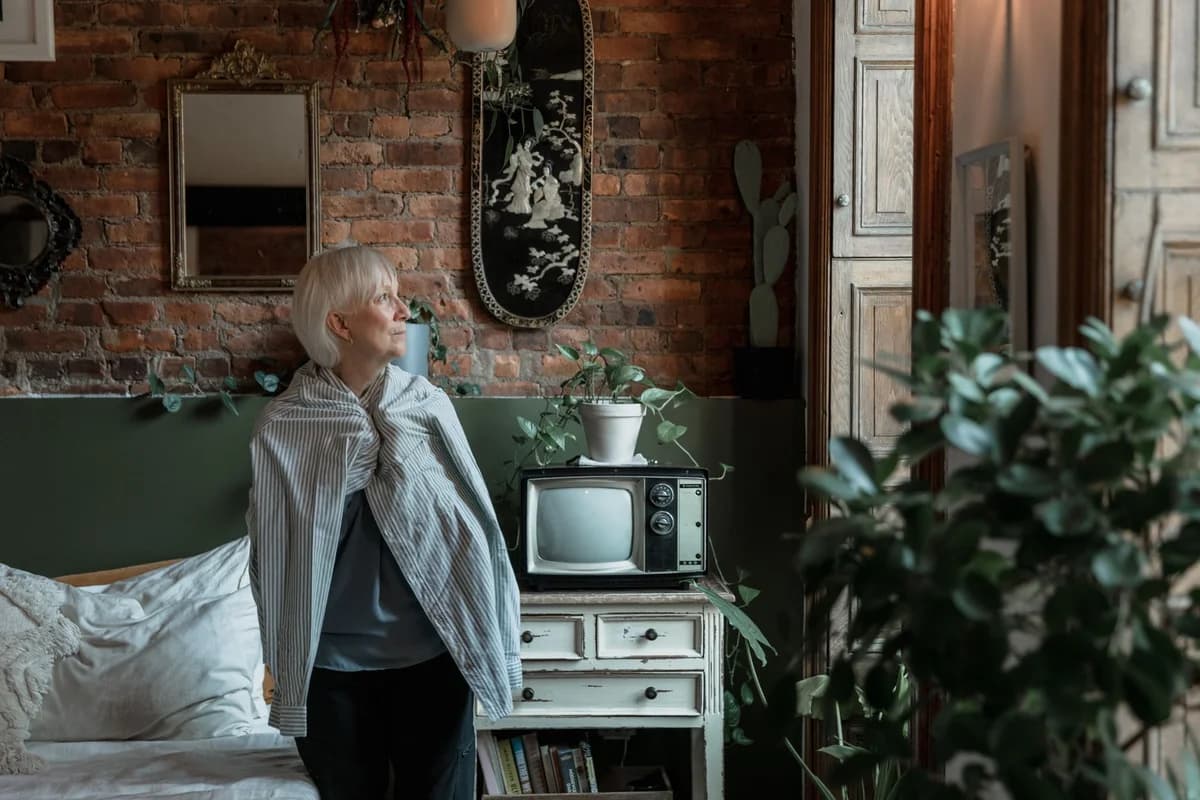 Elderly woman looks out window in cozy rustic room, surrounded by plants and vintage decor.