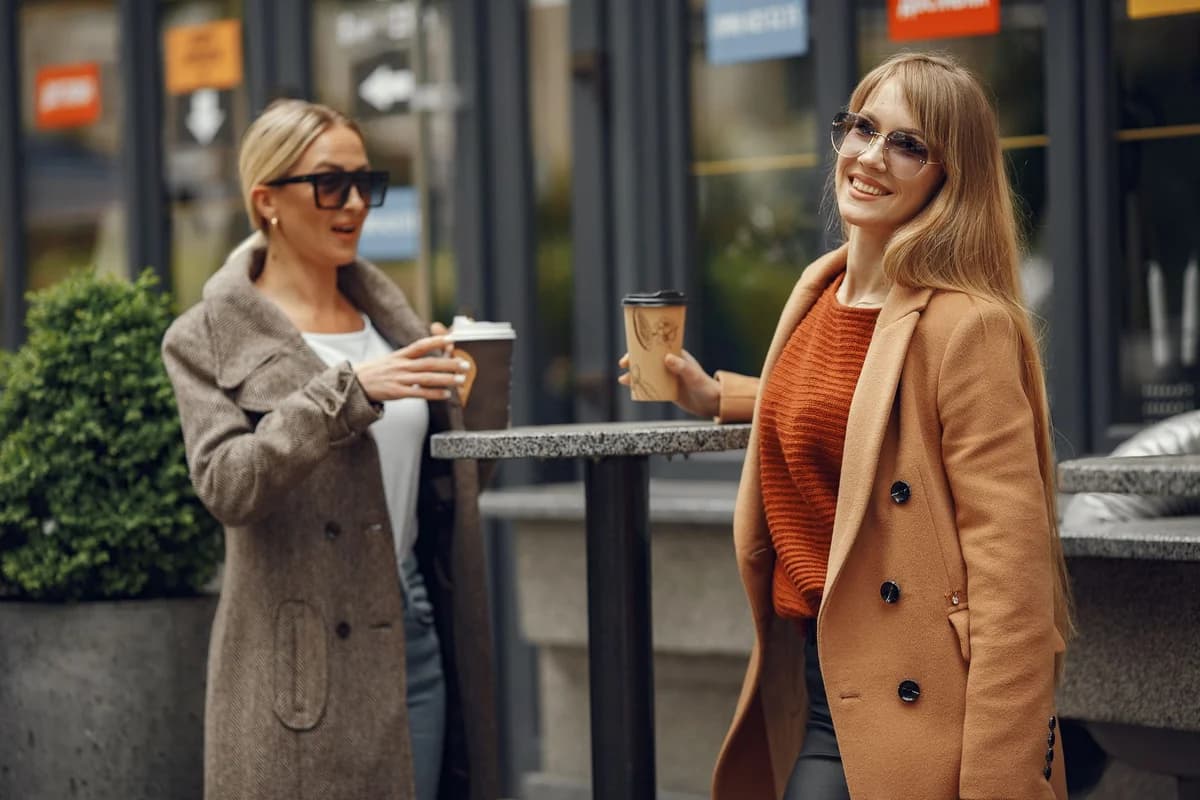 Two women in coats and sunglasses enjoying coffee and conversation on an urban street.