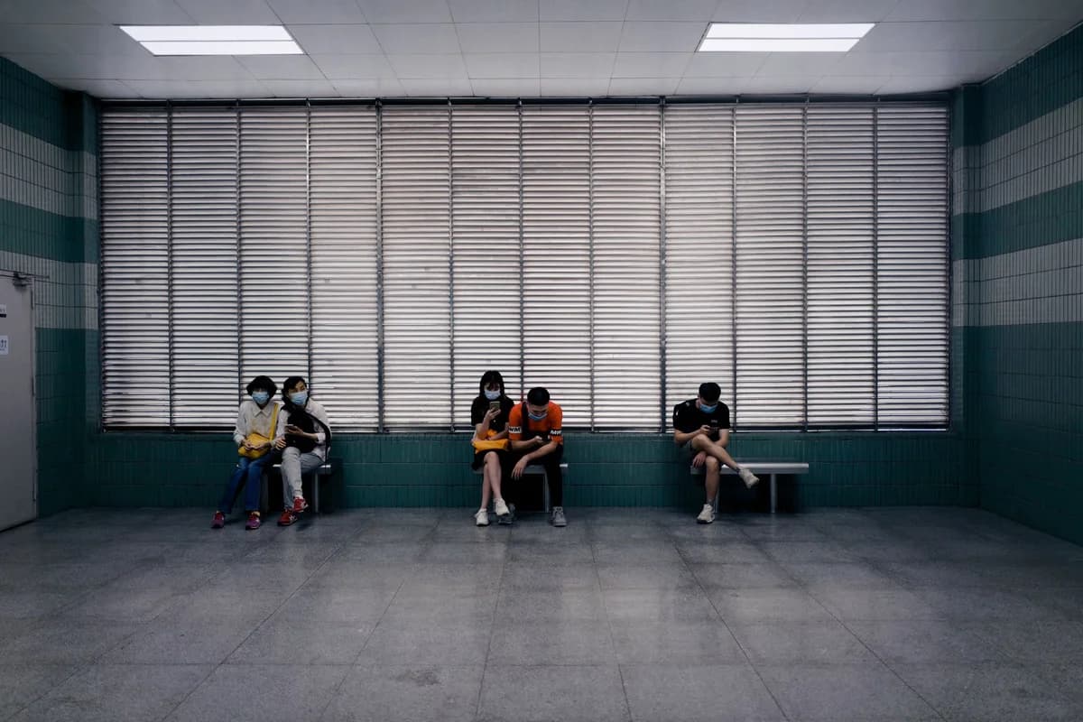 Full body of unrecognizable people in medical masks sitting on benches in spacious hall of clinic against windows