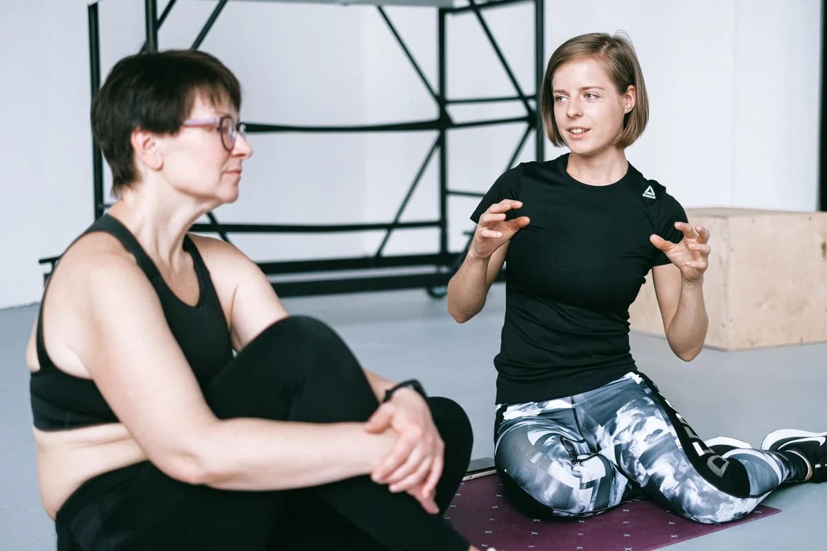Two women in sportswear sitting on a yoga mat, having a detailed discussion in a gym.