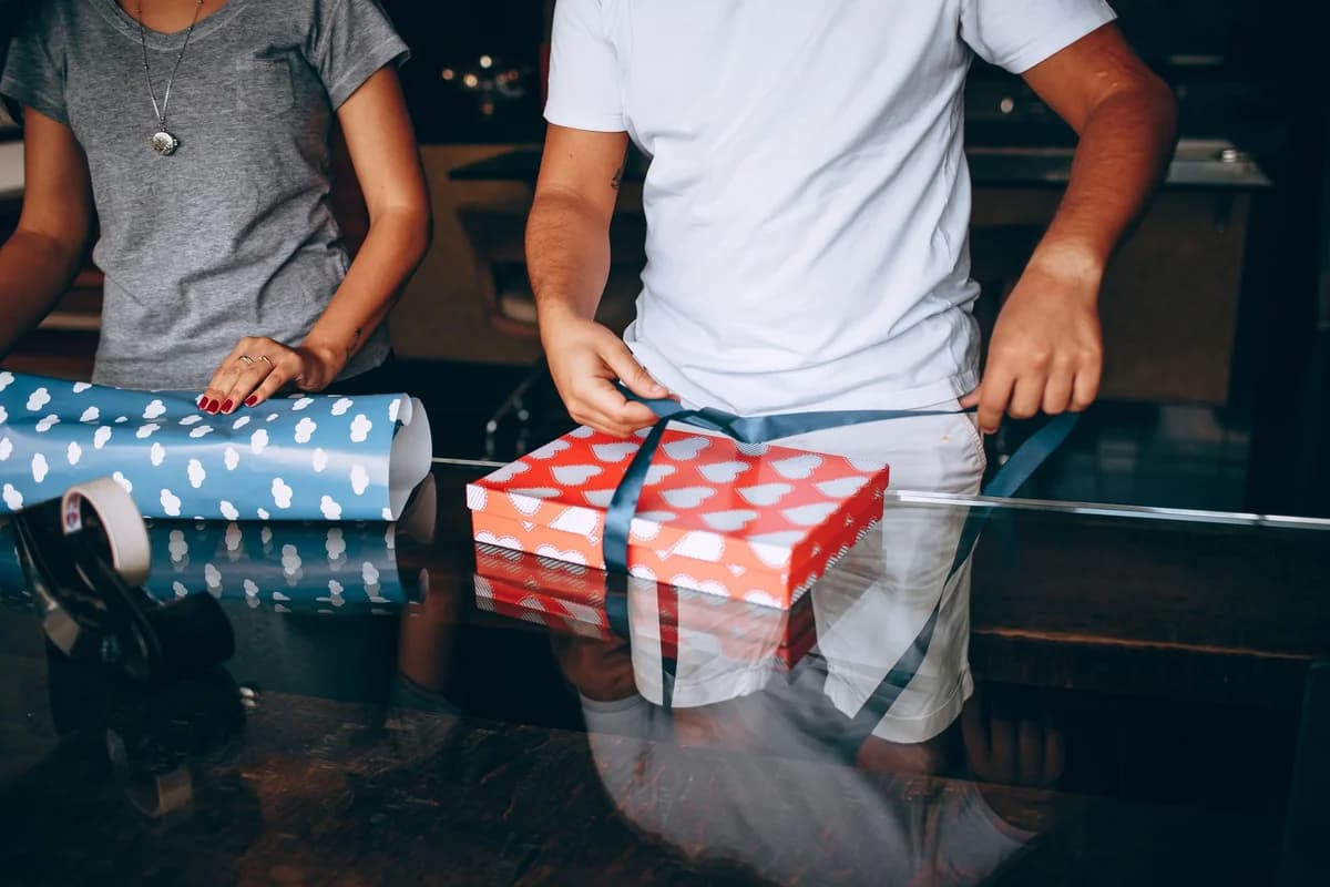 A man and woman wrapping a gift box with heart-patterned paper at home.