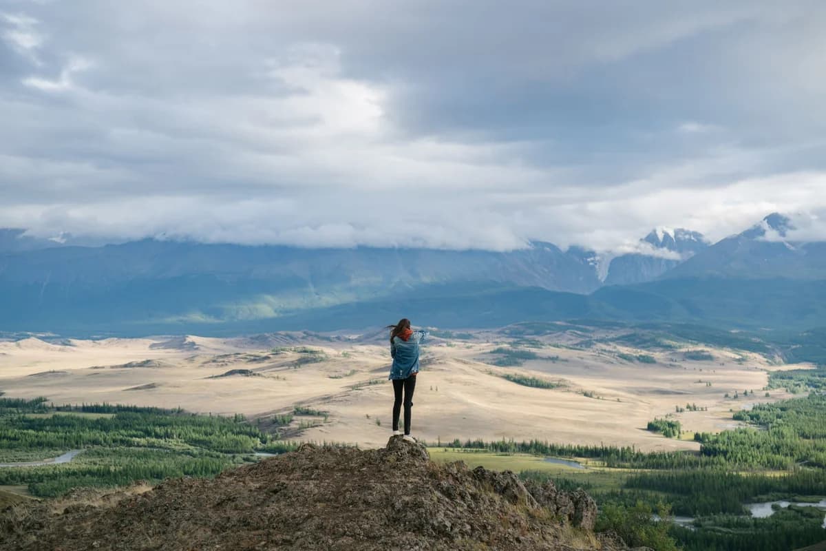 A person stands on a rocky peak surrounded by expansive mountain scenery under a cloudy sky.