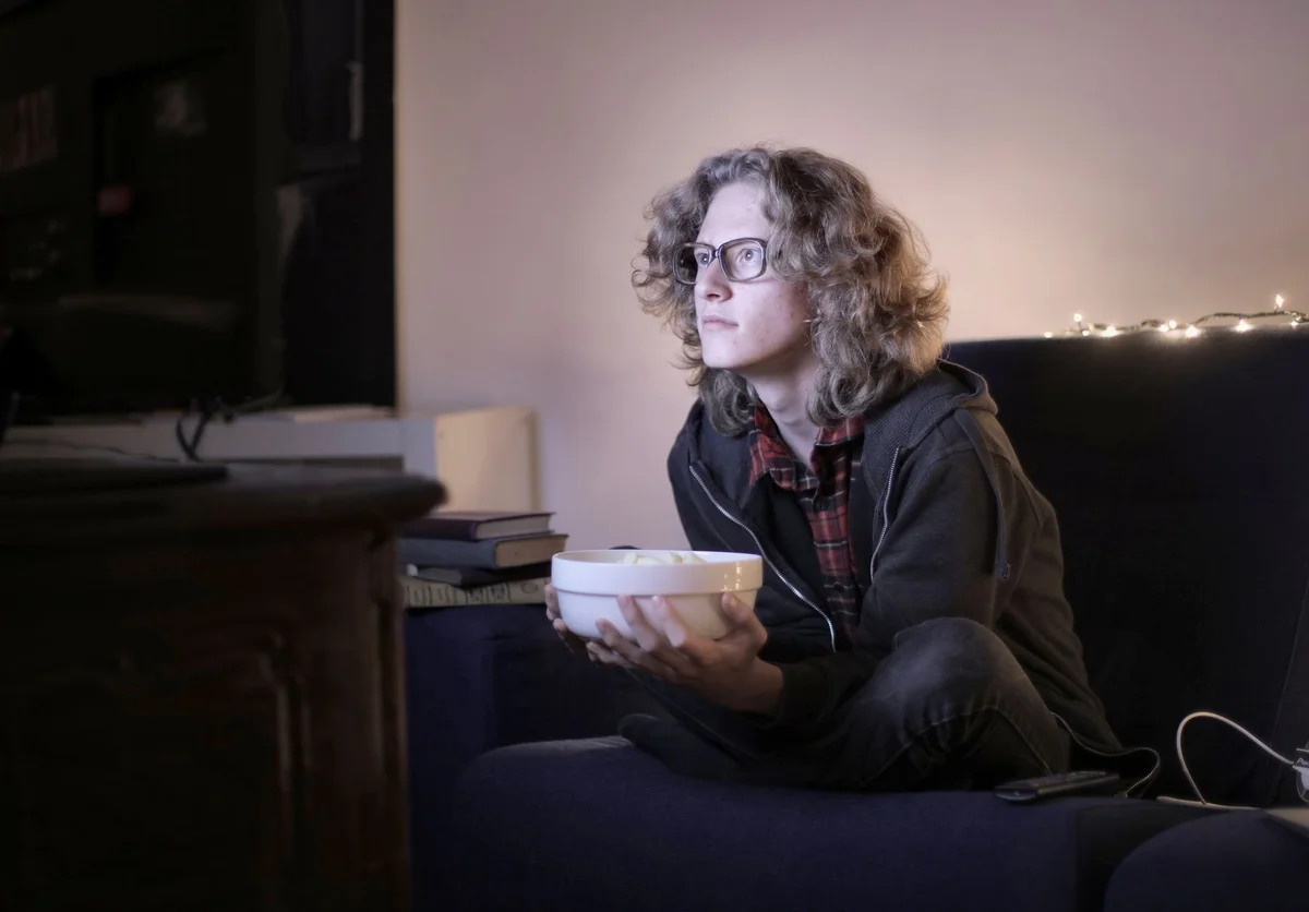 Concentrated male teenage with long hair in casual wear and eyeglasses sitting on couch with bowl of popcorn and watchin
