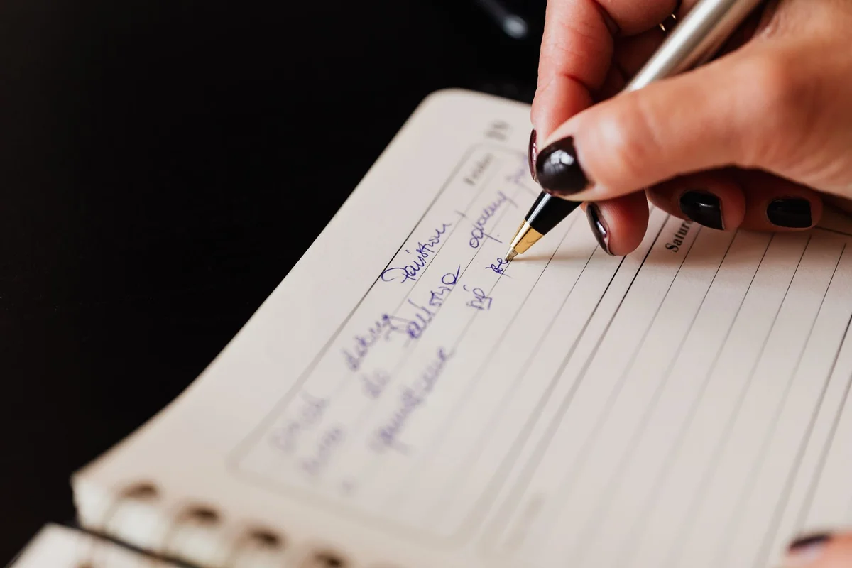 Closeup of crop anonymous lady with black manicure writing in planner with elegant pen