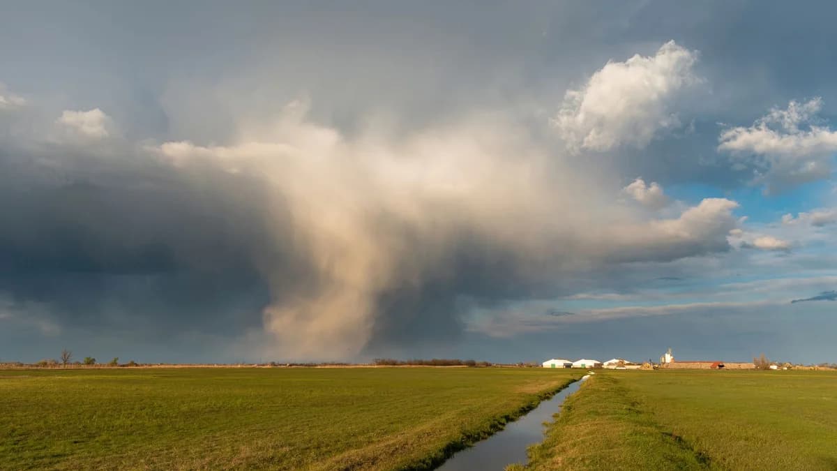 A dramatic cloud formation over a rural farm field with vivid green grass and a distant village.